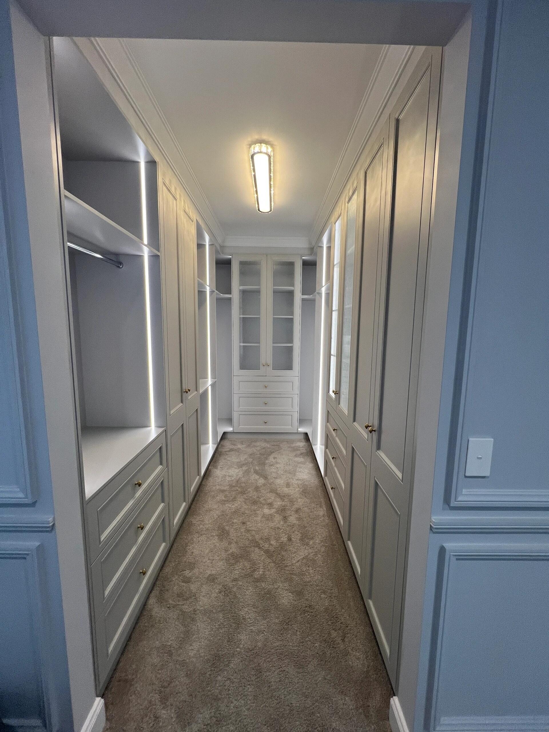 Walk-in wardrobe with white shaker doors, lit hanging space and a glass-door cabinet at the end