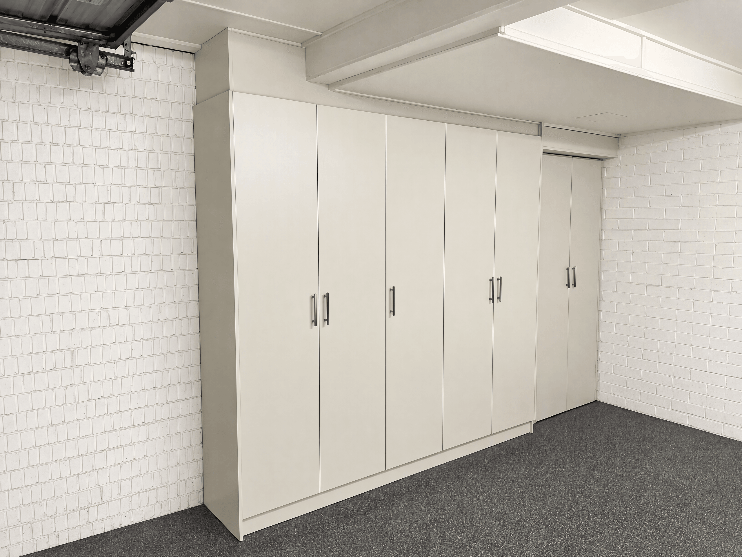 A run of tall white fitted storage cupboards with steel bar handles along a garage's brick wall