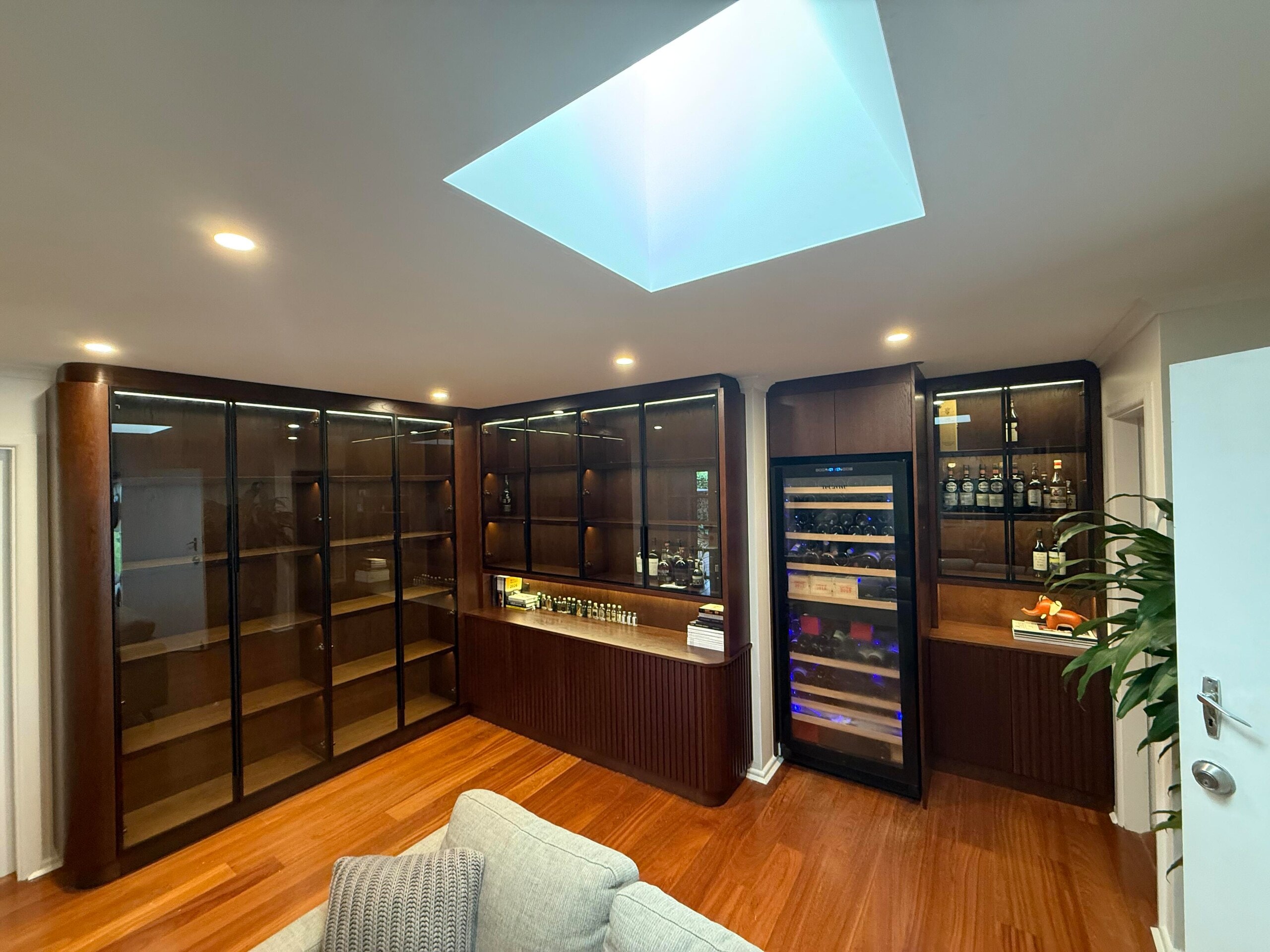 Dark timber bar wall with glass-door display cabinets, fluted lower doors and a built-in wine fridge
