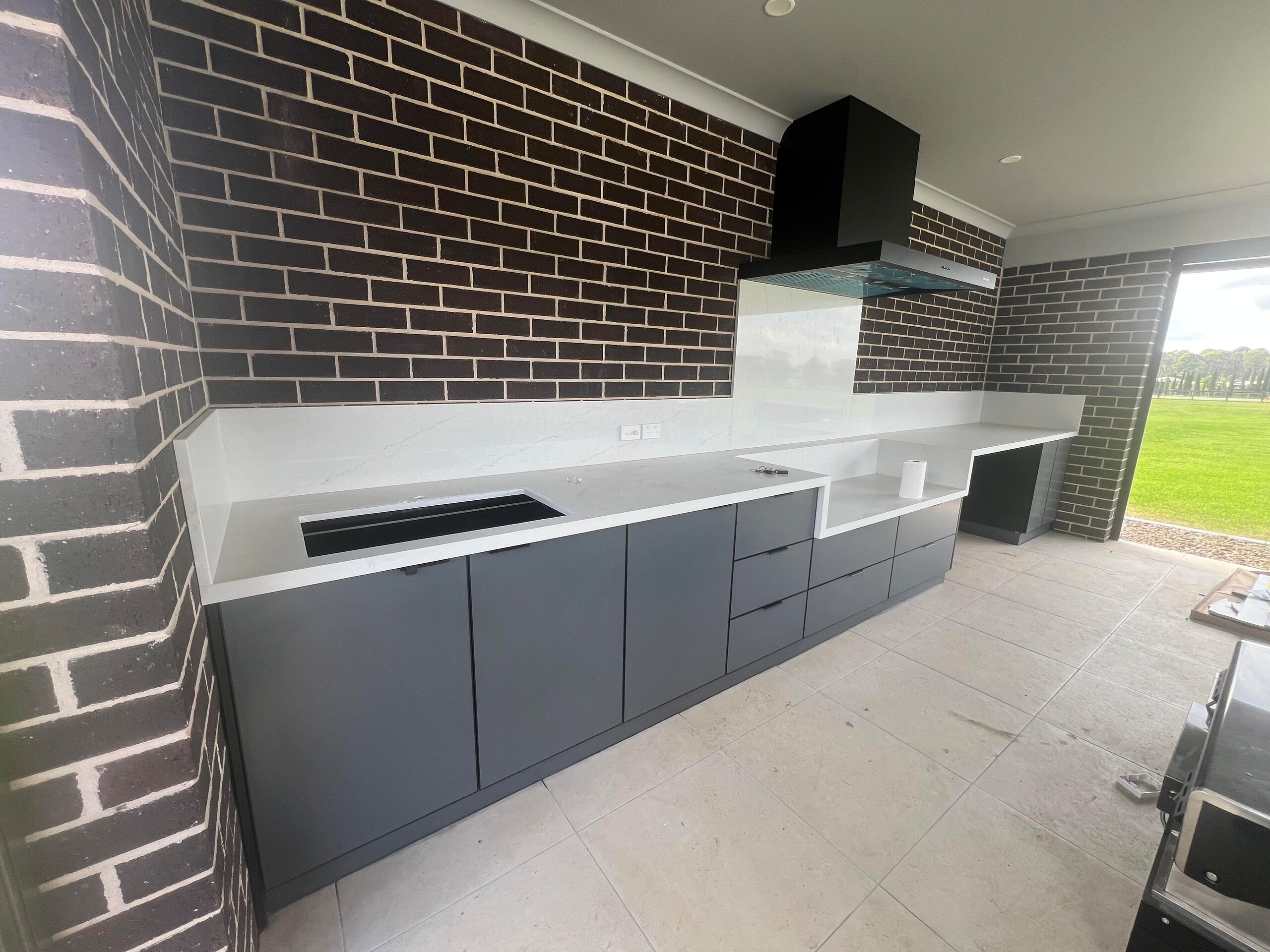Outdoor kitchen under a brick alfresco with charcoal cabinets and a stone-look benchtop